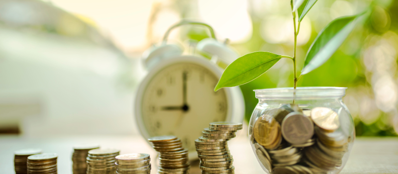 Coins stacked up and in a jar with a plant growing out of it. A white clock in the background
