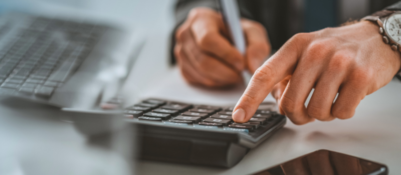 A person using a calculator with one hand and making notes with the other. A laptop in the background